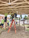Construction workers on ladders and scaffolding install wooden roof framing at a job site.