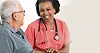 A smiling nurse talks with an elderly man in a bright indoor setting.