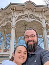Two people smiling for a selfie in front of a decorative white pavilion.