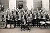 Large marching band standing in formation outside a stone building with drums and brass instruments.