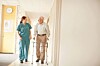 Healthcare worker and elderly man with a walker walking through a bright hallway.