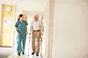 Healthcare worker and elderly man with a walker walking through a bright hallway.