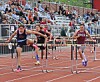 Three runners clear hurdles on an outdoor track in front of seated spectators.