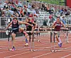Three runners clear hurdles on an outdoor track in front of seated spectators.