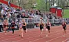 Several runners compete on a red outdoor track with spectators seated in the bleachers behind them.