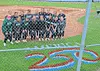 Youth baseball players pose for a group photo behind a chain-link fence on a baseball field.
