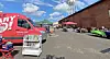 Outdoor market scene with vendor tables, umbrellas and a red promotional van beside a brick building.