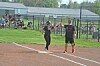Two people stand on a softball field near home plate with spectators seated behind a chain-link fence.