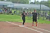 Two people stand on a softball field near home plate with spectators seated behind a chain-link fence.