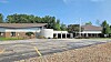 A low church building with a white cross, an empty parking lot, and trees under a blue sky.