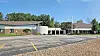 A low church building with a white cross, an empty parking lot, and trees under a blue sky.