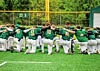 Youth baseball players in green uniforms huddle together on a grass field.