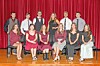 A group of people pose on chairs and standing in rows on a stage with red curtains behind them.
