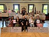 A group stands and sits indoors holding large check props in a room with flags and framed windows.