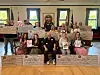 A group stands and sits indoors holding large check props in a room with flags and framed windows.