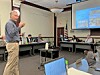 Man standing and speaking in a conference room with attendees seated at tables and a presentation screen visible.