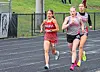 Three female runners racing on a wet track near a fence and grassy hill.