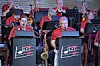 Jazz band members in red shirts play brass instruments on stage behind branded music stands.