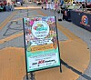 Farmers market sign on a sidewalk with vendor booths and people in the background.