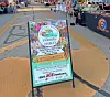 Farmers market sign on a sidewalk with vendor booths and people in the background.