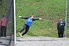 Athlete in blue extends arms while standing in a discus throwing cage with an official nearby.