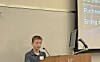 A student speaks at a podium in front of a classroom projection screen.