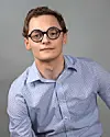 Portrait of a man in round glasses and a light blue shirt against a gray background.