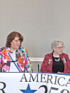 Two women stand behind a banner reading America 250 at a public event.