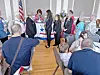 People gathered in a room around a podium with a star logo, with U.S. flags visible behind them.