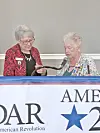 Two women stand behind a banner at an indoor event, one holding a microphone.
