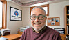 Smiling man wearing glasses sits in an office with a People’s Voice logo frame on the wall.
