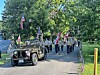 People march behind a vintage jeep while carrying American flags and a patriotic banner on a road lined with trees.