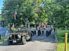 People march behind a vintage jeep while carrying American flags and a patriotic banner on a road lined with trees.
