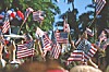 Crowd waving small American flags outdoors near trees and red, white and blue balloons.