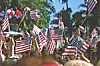 Crowd waving small American flags outdoors near trees and red, white and blue balloons.