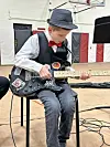 A boy in a fedora and bow tie plays an electric guitar while seated indoors.