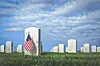 Rows of white military headstones with American flags in a grassy cemetery under clouds.