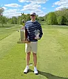 Golfer standing on a fairway holding two trophies on a sunny golf course.