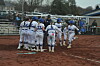 A softball team in white uniforms huddles on a dirt field with spectators in the stands behind them.