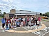 A large group of people poses outside a single-story building with a parking lot and cloudy sky.