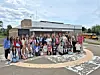 A large group of people poses outside a single-story building with a parking lot and cloudy sky.