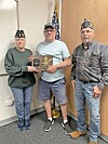 Three veterans standing indoors, with two holding a commemorative plaque in front of a U.S. flag.