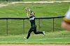 A softball player jumps with a glove raised to catch a ball near the outfield fence.