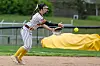 Softball player in a striped uniform reaches low for a ball on a dirt infield.