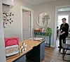 Small reception room with desk, welcome sign, wall mirror, plants, and chairs, with a person standing near the door.