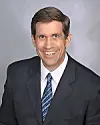 Studio portrait of a smiling man in a dark suit, white shirt and blue tie on a gray background.