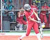 Softball player in red swings at a yellow ball near home plate with teammates watching from the dugout.