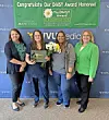 Four women stand in front of a VVMedica backdrop, with one holding a Daisy Award plaque and flowers.