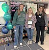 Three women pose in a classroom beside teal and white balloons and a decorated bulletin board.