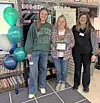 Three women pose in a classroom beside teal and white balloons and a decorated bulletin board.